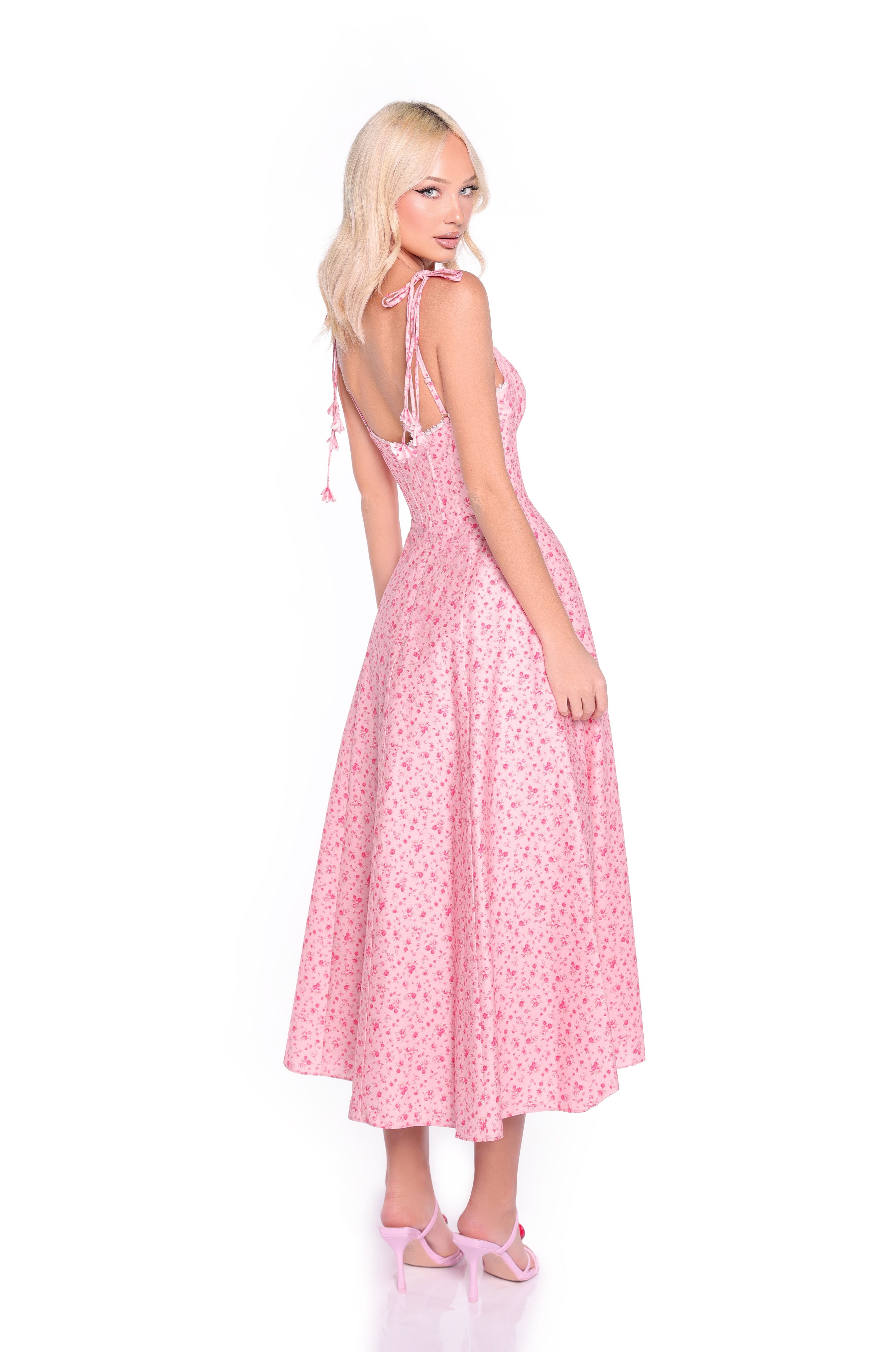 A woman models the Casual Midi Pink Floral Dress: Aria, featuring a light pink floral print, corset bodice, and tie shoulder straps. She stands against a white background, shown from the side and back.